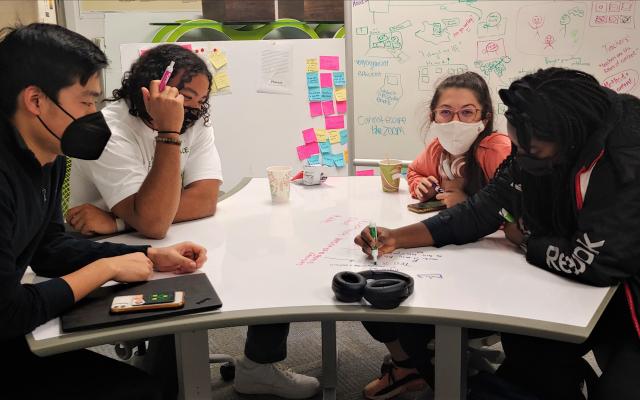 2 male students on the left, 2 female students on the right, sitting around a dry-erase desk